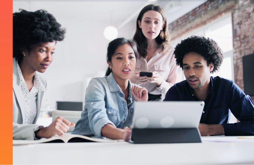 Three people sitting at a table while they all look at a tablet device while another person is standing behind and looking as well