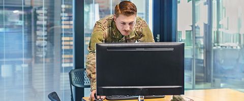 A person in camouflage clothing leaning over a computer monitor while using a mouse to view something on the screen 