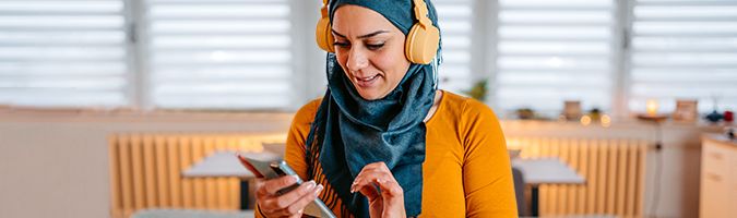 Woman in a hijab sitting at a table while listening to headphones and looking at a tablet device