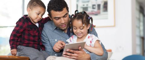 An adult and two children look at a tablet the adult is holding with artwork on the walls in the background