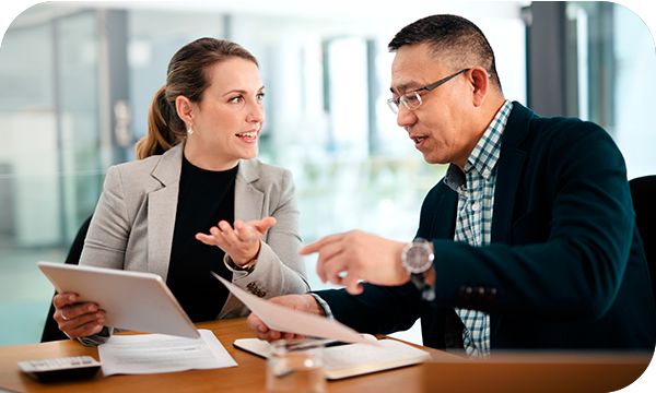 Two coworkers sit together at a desk and discuss information on a sheet of paper and tablet in their hands