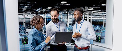 Three individuals in a manufacturing facility discussing important data on a laptop while one marks a checklist on a clipboard.