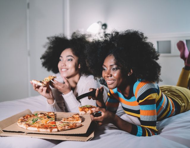 Two women laying on a bed while eating pizza and watching TV
