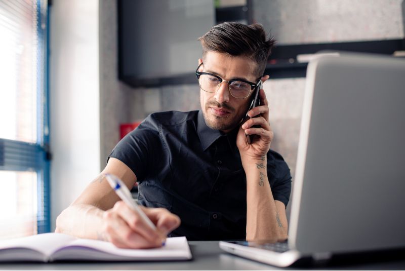 A person sits at a desk in front of a laptop while taking notes and talking on a cell phone. 