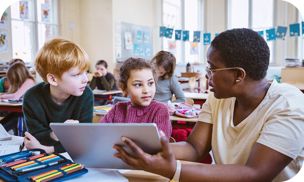 An educator helps two students with a tablet in a classroom setting as other students work at tables in the background