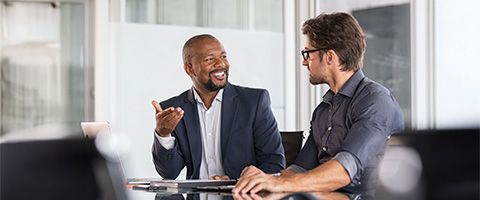 Two people in business attire smile and talk in front of a window.