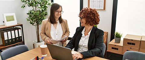 Two people in business casual attire standing and sitting around an office table with a laptop. 