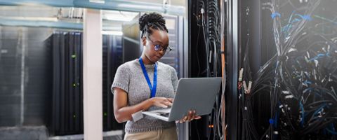 A person standing in a server room reviews information on a laptop 