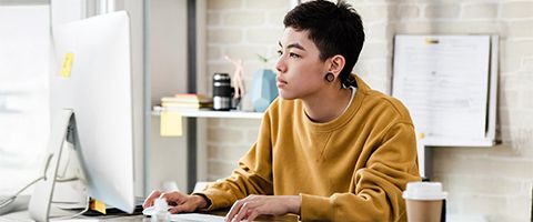 Person in a mustard-colored sweater working at a desk with a desktop computer, coffee cup and office supplies in the background.