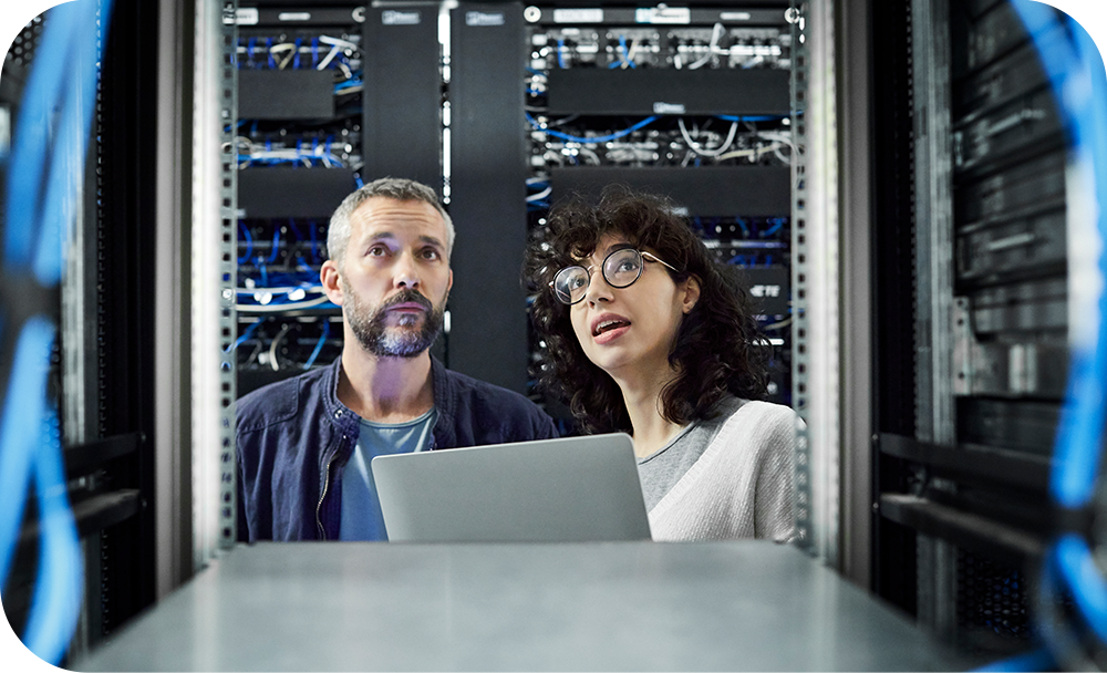 Two people in a server room with a laptop are surrounded by racks of servers and cables