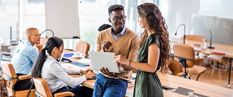 Two people standing and talking to each other as one holds an open laptop with people at desks in the background