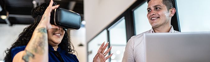 Woman wearing a VR headset while standing next to a man in front of a laptop