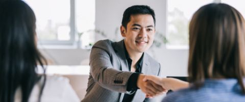 A smiling businessperson in a grey suit reaches across a desk to shake the hands of two other businesspeople 