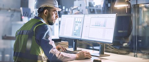 A worker in an energy facility wearing a hard hat and safety vest looks at two computer monitors 