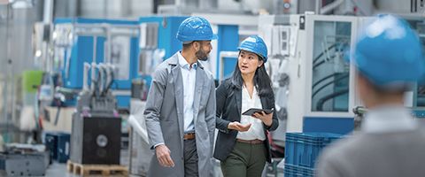 Two employees wearing hard hats talk while walking through a power plant