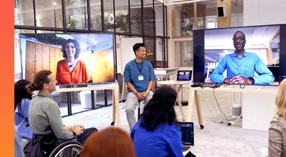  A man sitting on a stool in front of an audience near two large monitors with video call participants