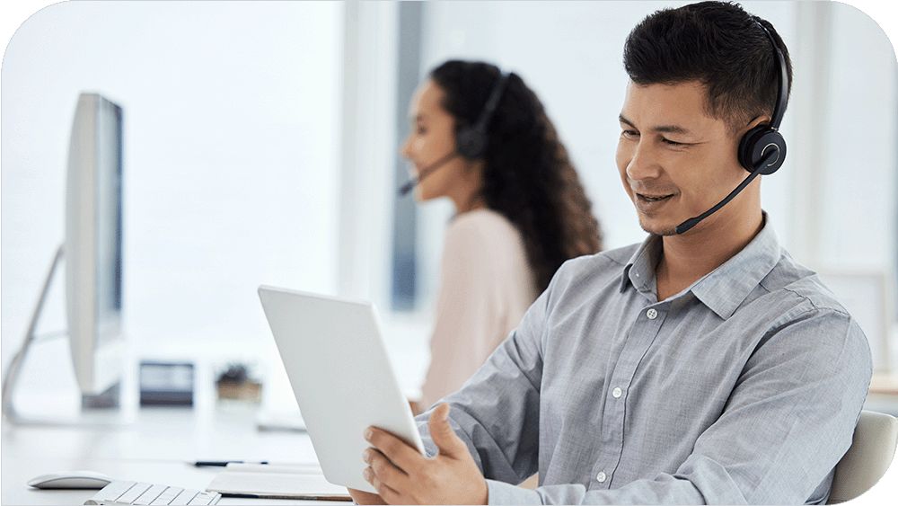  A man wearing a headset works on a tablet while a woman works in the background in a bright office