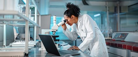 A person wearing a lab coat looks at a microscope and records results on a laptop.  