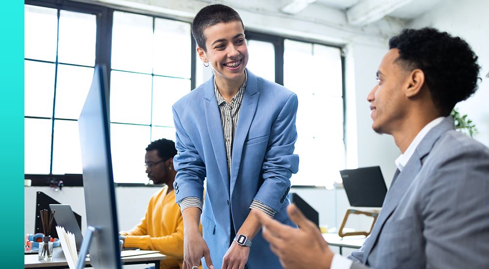 Person in a light blue suit jacket standing while talking to another person sitting at a desk in front of a computer monitor