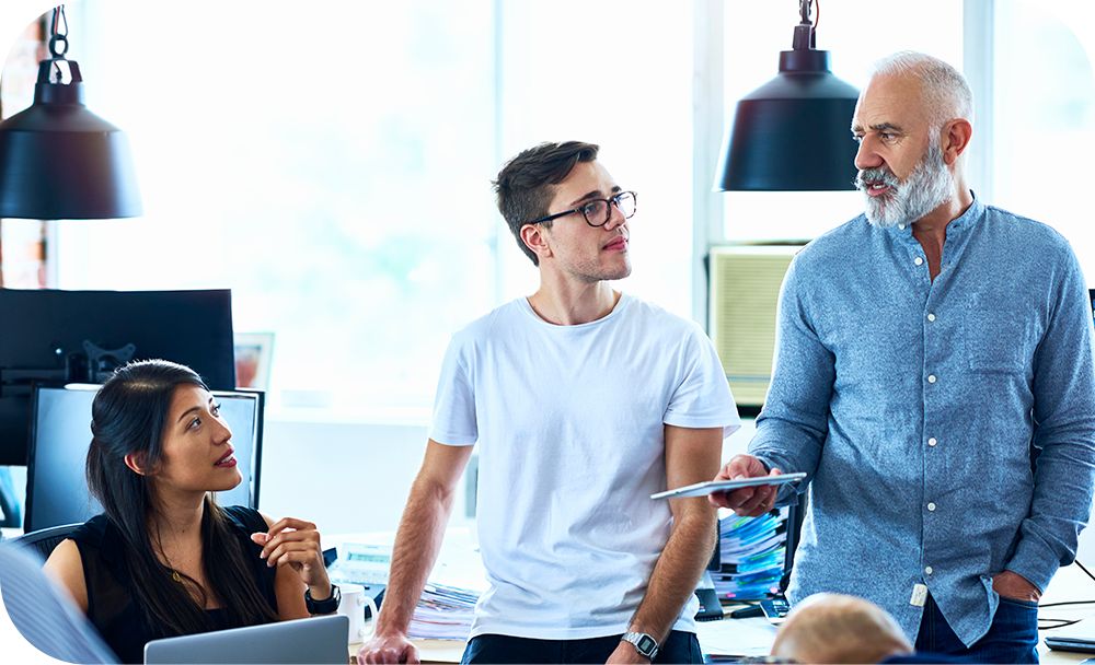 Woman sitting in front of a desktop computer while talking to two men standing next to her while one holds a tablet device