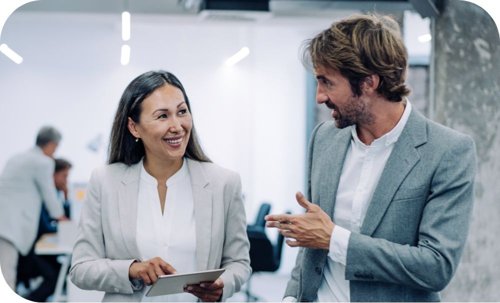 Two business professionals wearing suits are engaged in a discussion in a modern office, one holding a tablet