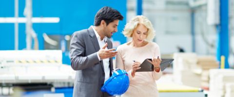 Two professionals discussing work, one holding a blue helmet and the other holding a tablet, in an industrial setting