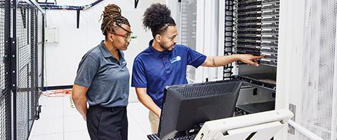  Two colleagues in server room examining data on computer monitor on desk in front of them.