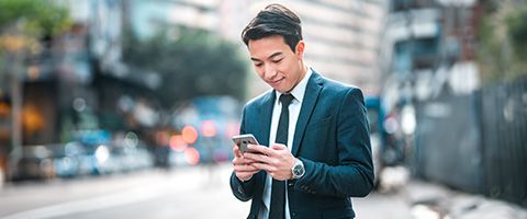 An individual in a business suit holds and looks at a mobile phone with a busy urban setting in the background