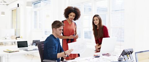 Man sitting down and two woman standing looking at paper at a desk