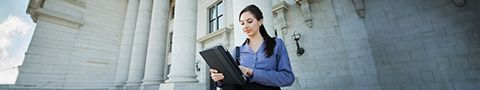 A businessperson in a blue button-up sits outside on the steps of a federal building working on their tablet