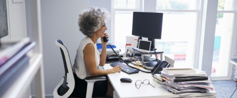 Businessperson at a desk talking on the phone and looking at two computer screens with papers, a notebook, glasses and a mobile phone on the desk