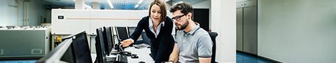 A woman discusses something on a man’s monitor screen.