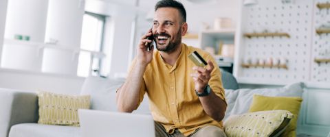 A person holds a credit card and talks on the phone in front of a laptop while seated on a couch