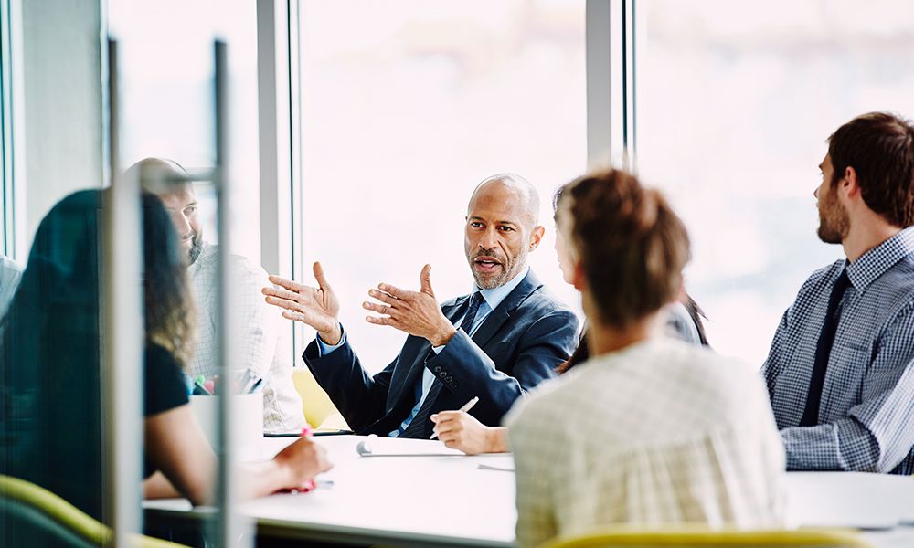 Six people in business attire sitting at a conference table having a discussion in an office setting. 