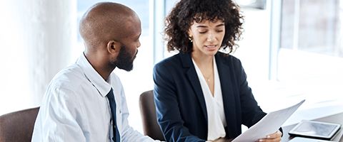 Two people in business attire sitting at a table and talking while looking at a piece of paper. 