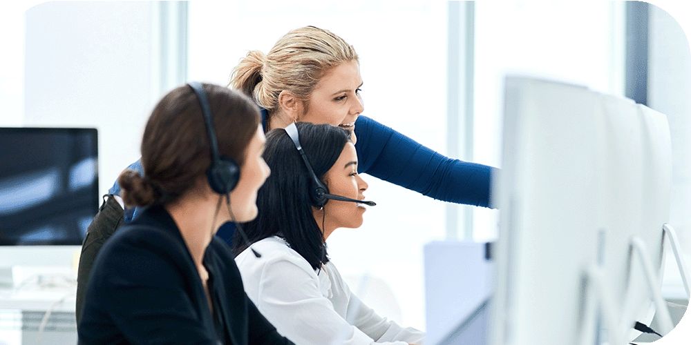 Two call center workers wearing headsets sit at their computers while a person standing next to them points at something on the screen