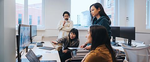 Four people in an office sit looking at information on a computer screen as one of them gestures to it