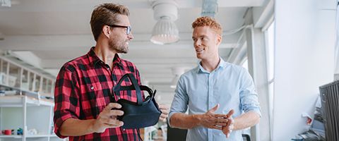 A person holds a VR headset while standing and talking with another person