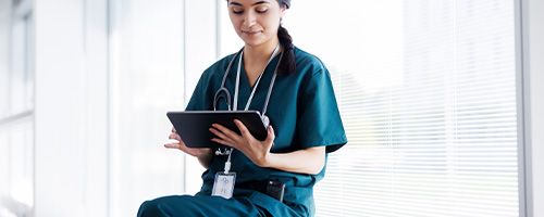 Woman in nursing uniform sitting down while working on a tablet device