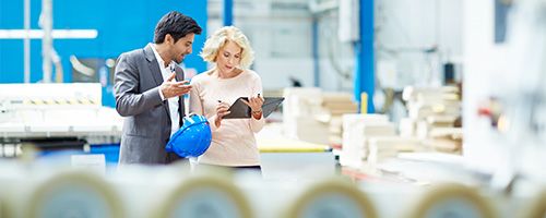 Woman holding a tablet device standing next to a man holding a blue hard hat on a factory floor