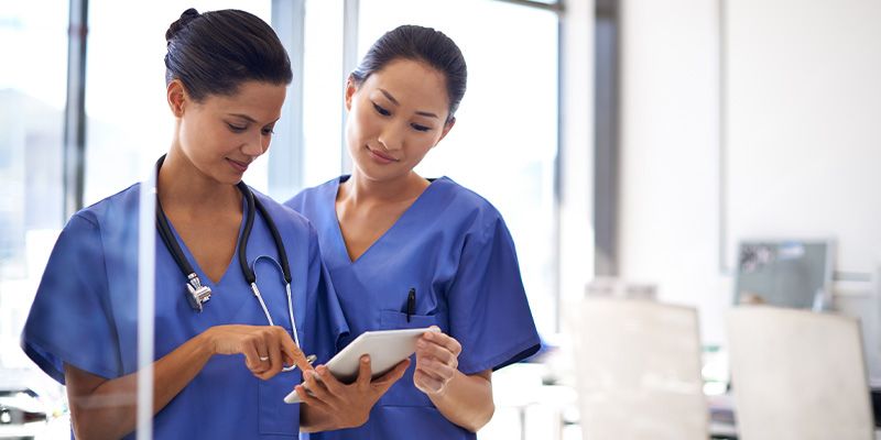 Two women in nursing uniforms standing while looking intently at a tablet device