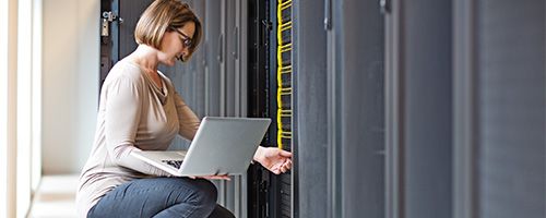 Woman kneeling in front of a server stack while holding a laptop on her knee