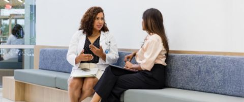 A woman in a white lab coat sits on a bench in an office lobby and talks to a professional woman sitting next to her