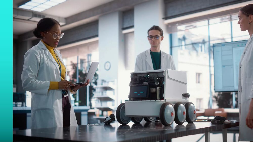 Three people in white lab coats observe a robotic instrument with six wheels on a table in front of them