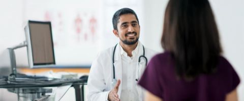 A smiling medical doctor with a stethoscope around his shoulders talks to a women seated across from him in a medical office 