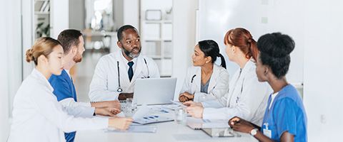 A group of medical professionals sits around a table reviewing patient cases on a laptop 
