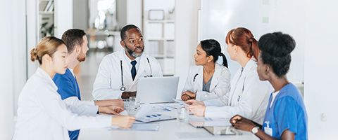 A group of medical professionals sits around a table reviewing patient cases on a laptop 