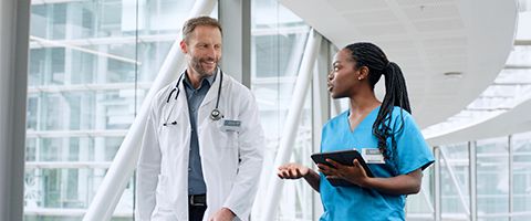 A medical professional wearing hospital scrubs holds a tablet and walks down a hallway talking to a person wearing a lab coat and stethoscope