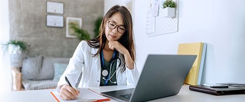 Person wearing a lab coat sits at a desk viewing information on a laptop while taking notes 