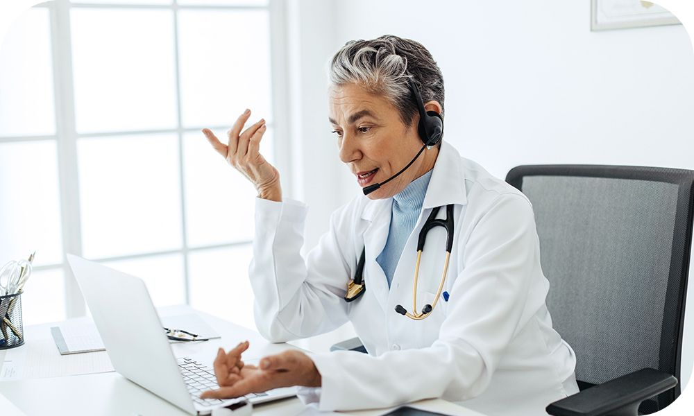 A doctor sitting at a desk in front of an open laptop gestures while participating in a video conference call